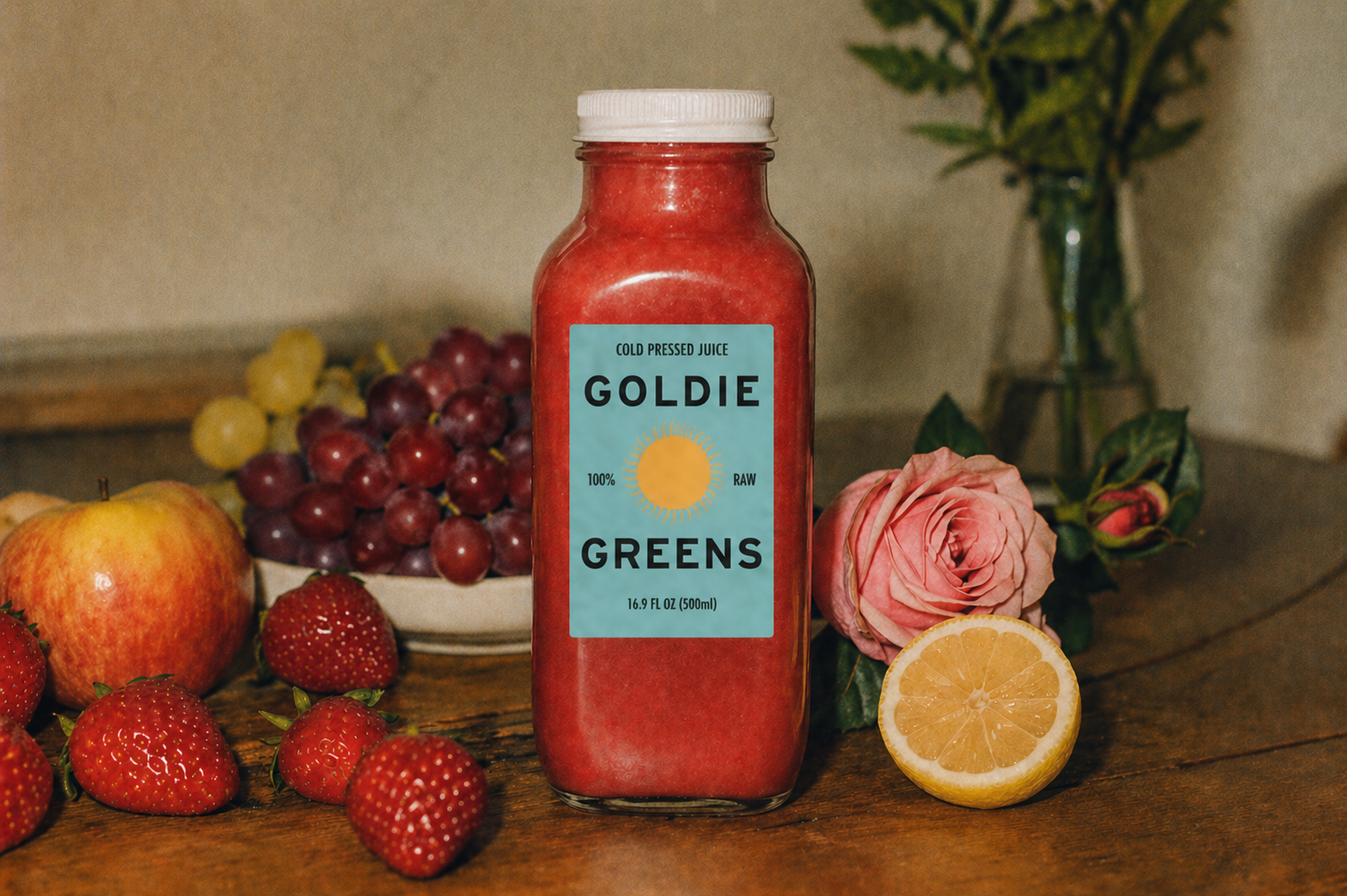 A bottle of Strawberry Fields cold-pressed juice on a wooden table, surrounded by fresh strawberries, grapes, apples, a rose, and a halved lemon.